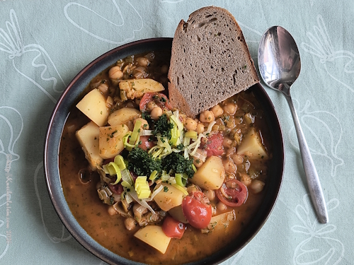 Teller mit Eintopf mit Kichererbsen, Kartoffeln und Tomaten mit einer Scheibe Brot, daneben ein Esslöffel und e