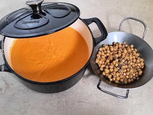 Cremige Karottensuppe mit Tahini und Rosmarin nach Anna Jones im Topf, daneben eine Pfanne mit gerösteten Kichererbsen