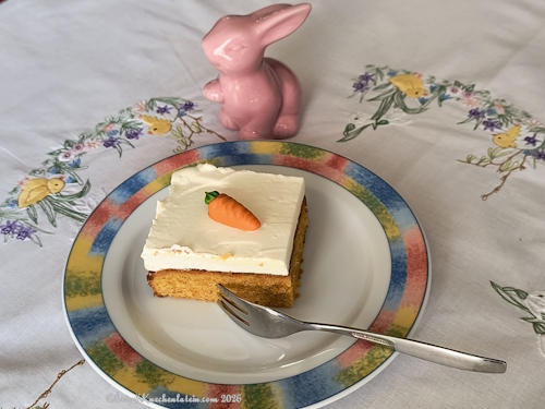 Ein Anschnitt von Schwedischer Möhrenkuchen - Morotsrutor Goldbrauner Teig mit sichtbaren Karottenstücken unter einer dicken Schicht weißer Creme dekoriert mit einer Marzipanrübe