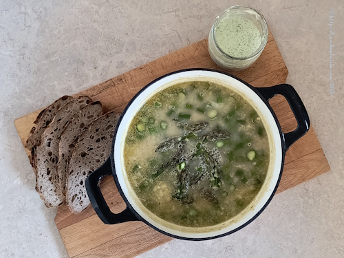 Topf mit frisch gekochter Spargelsuppe mit Graupen und gelben Erbsen, daneben ein Glas mit Petersiliencreme und knusprige Brotscheiben