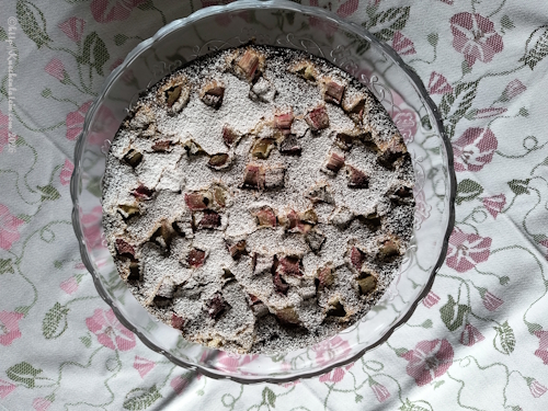 Rhabarber-Kladdkaka mit weißer Schokolade und Kardamom mit Puderzucker bestreut auf Glastortenplatte auf Tischdecke ÅKERVINDA