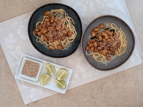 2 Teller Pasta mit karamellisierten Bohnen und Spinat, daneben 1  Schälchen gerösteter  Sesam und Limettenviertel auf Porzellan